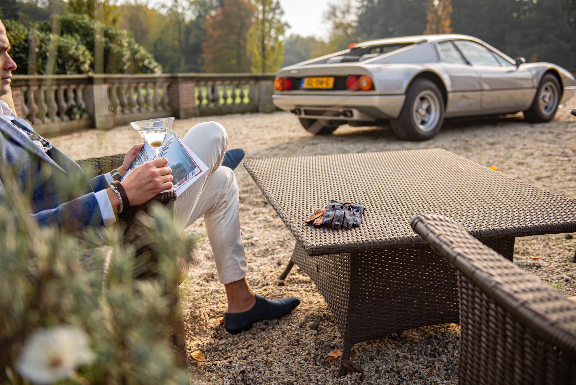 A man with blue driving gloves, in a blue blazer and cream trousers sits outdoors holding a drink and magazine, with a brown wicker coffee table in front of him and a silver Ferrari classic sports car parked on a gravel driveway in the background.