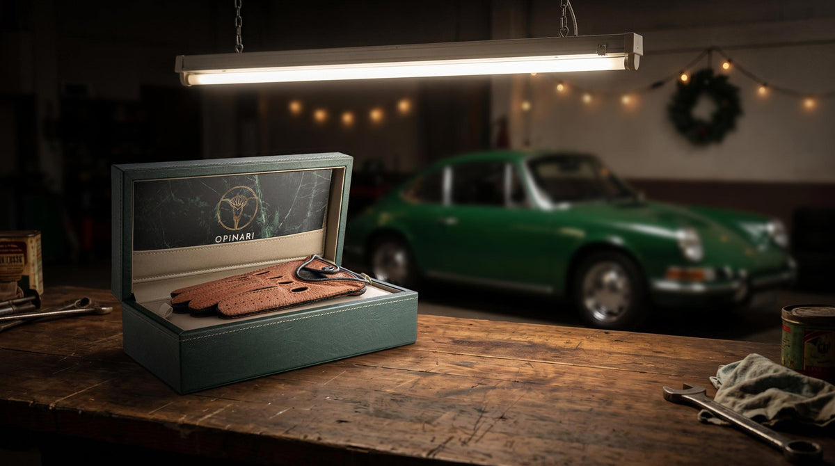 A teal presentation box containing a pair of brown leather gloves, displayed on a rustic wooden workbench in a garage setting with a green classic car and warm hanging lights blurred in the background.