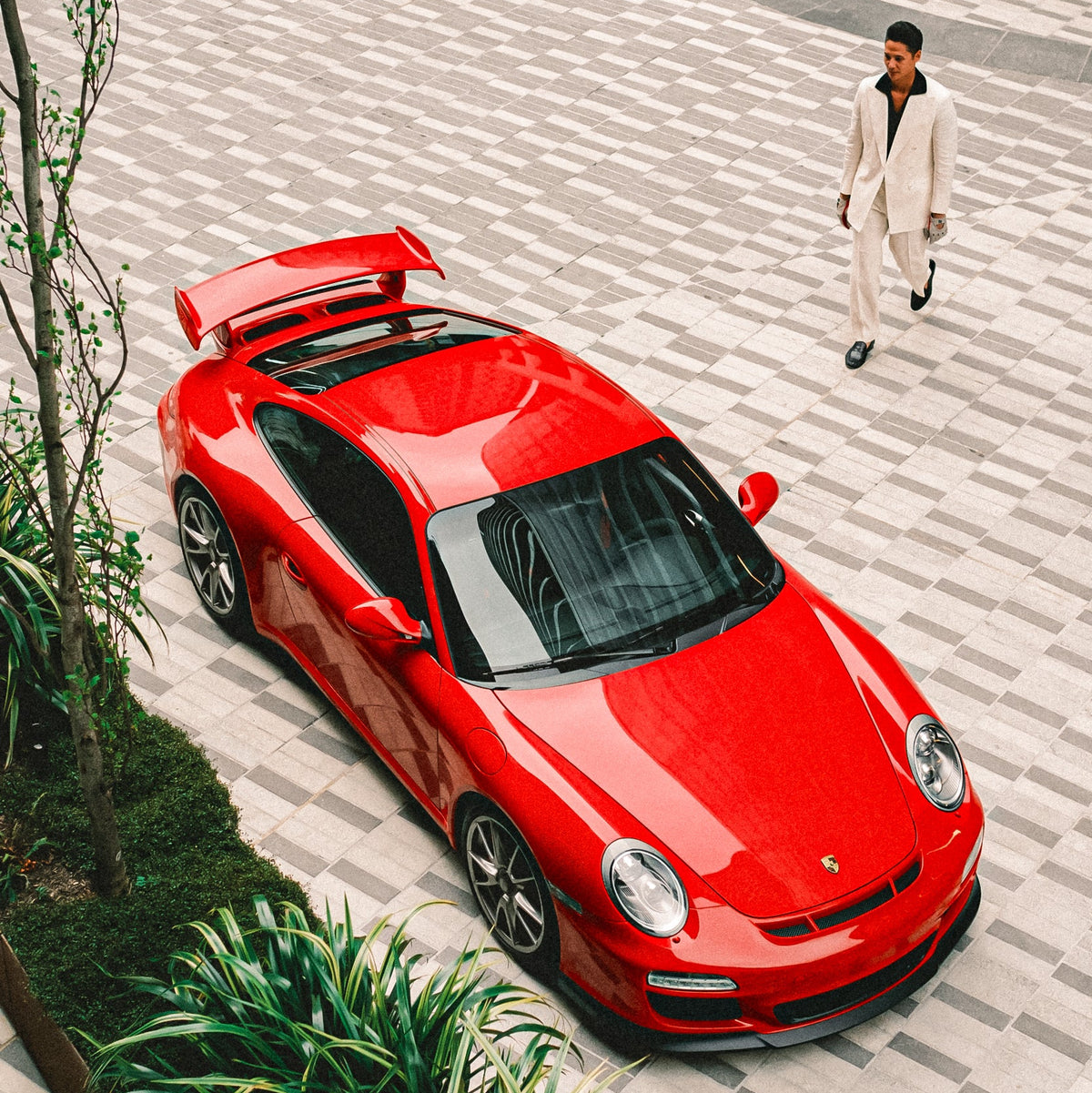 A red Porsche sports car is photographed from an elevated angle on a paved plaza, with a man in a light-colored suit walking nearby and green plants along the left side.