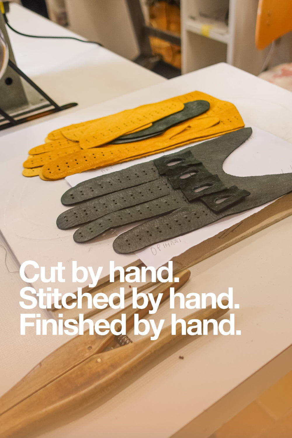 A pair of handmade leather gloves, one yellow and one dark gray, laid out on a white table with a wooden tool. White text overlay reads: 'Cut by hand.