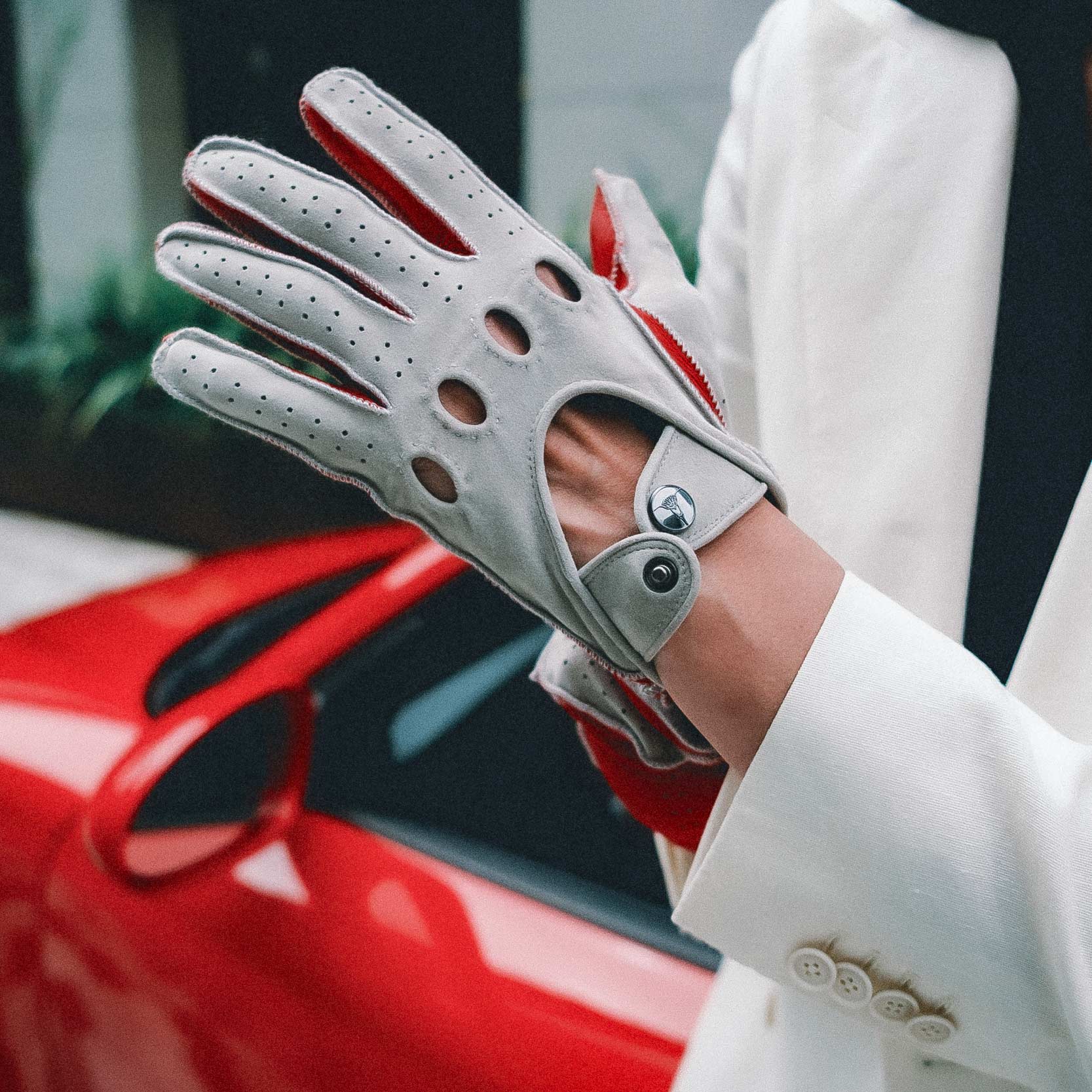 Gray driving glove with red accents held by a person in a white suit against a blurred red car background