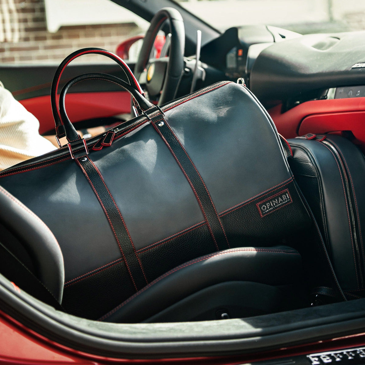 A black leather D'Amore duffel bag with red contrast stitching and handles, resting inside a red and black sports car.
